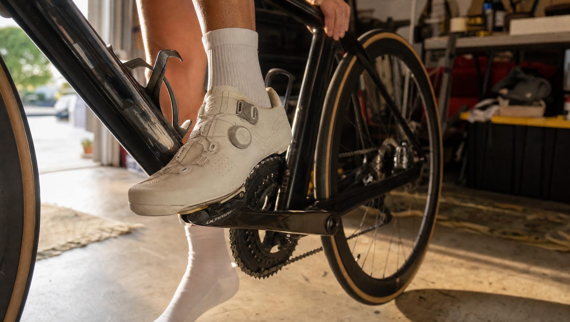 A cyclist clipping a cycling shoe into a road bike pedal in a sunlit home garage.