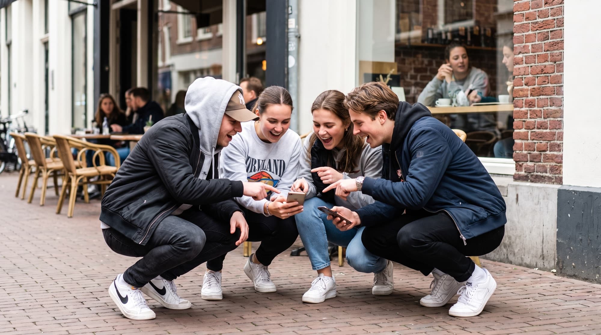 A group of friends crouched together outside a cafe, laughing and comparing something on their phones.